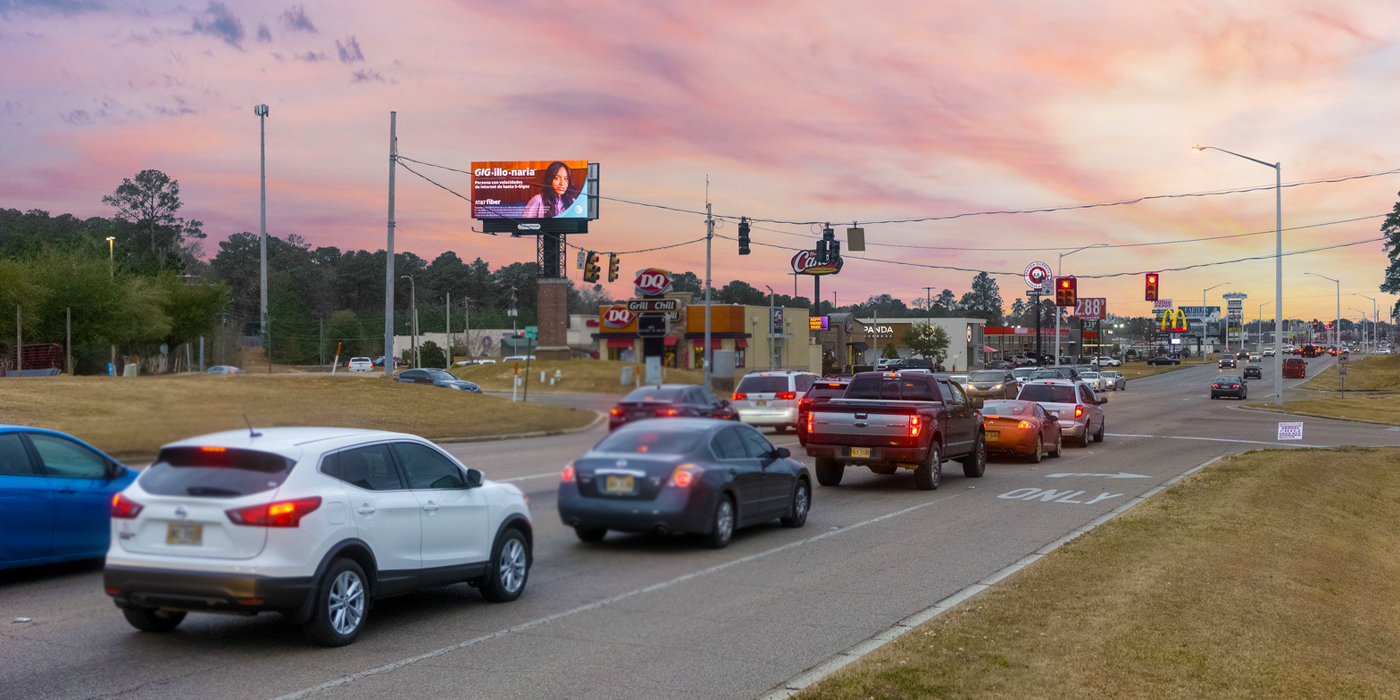 Cars driving under a large digital billboard on a highway during sunset.