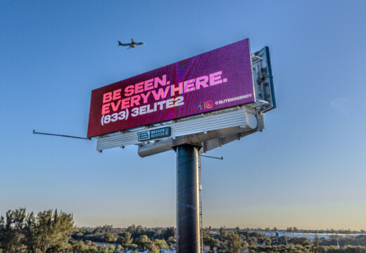 Large pink digital billboard reading “Be Seen. Everywhere.” with airplane flying overhead.