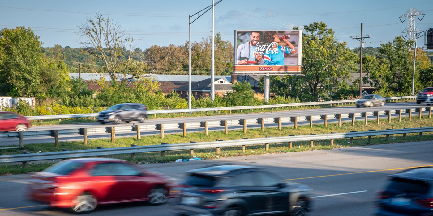 Vehicles passing by a tall digital billboard with a blank screen near trees and sky.