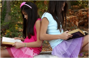 Two young girls sitting back-to-back outdoors reading