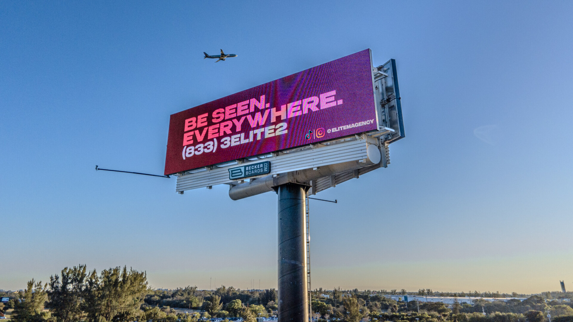 Large pink digital billboard reading “Be Seen. Everywhere.” with airplane flying overhead.