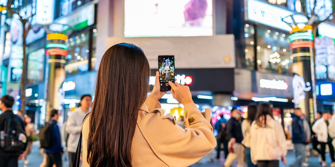 Woman photographing large digital billboards in a city square at night.