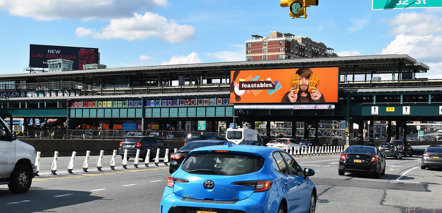 Bright orange digital billboard advertising Feastables chocolate brand with man holding chocolate bars near busy city traffic.