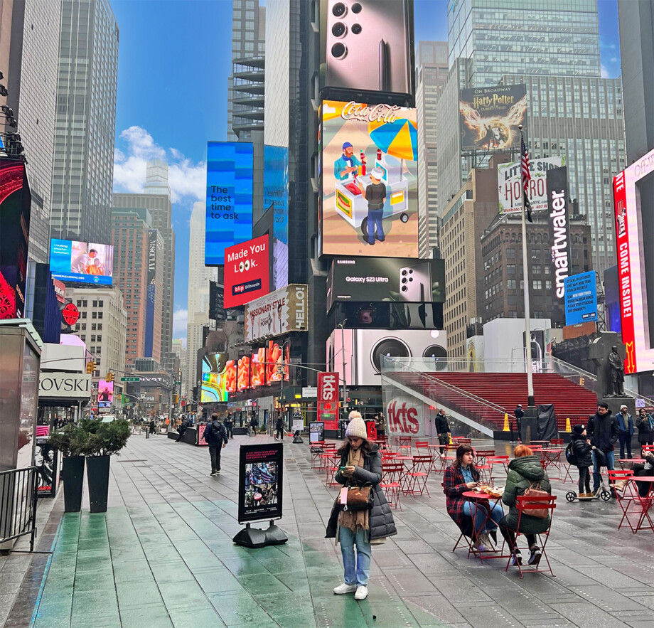 Busy Times Square street with large colorful digital billboards, pedestrians walking across crosswalks.